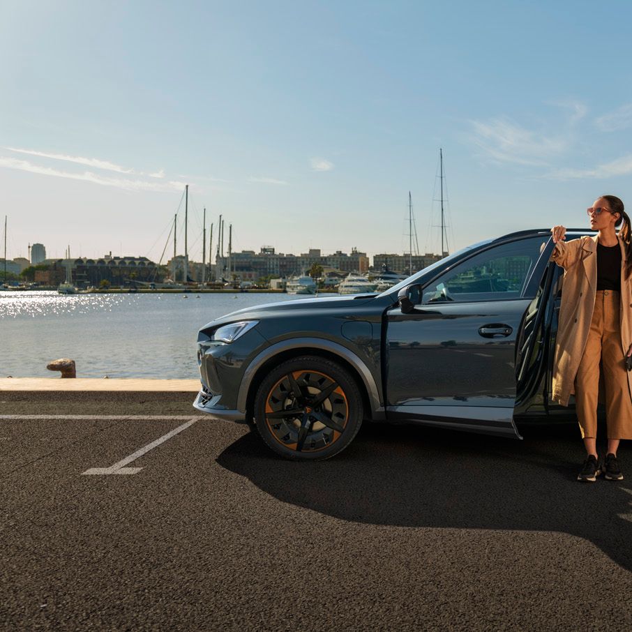 Eine Frau steht an der geöffneten Vordertüre eines grauen CUPRA Leon der am Kai eines Hafens parkt.