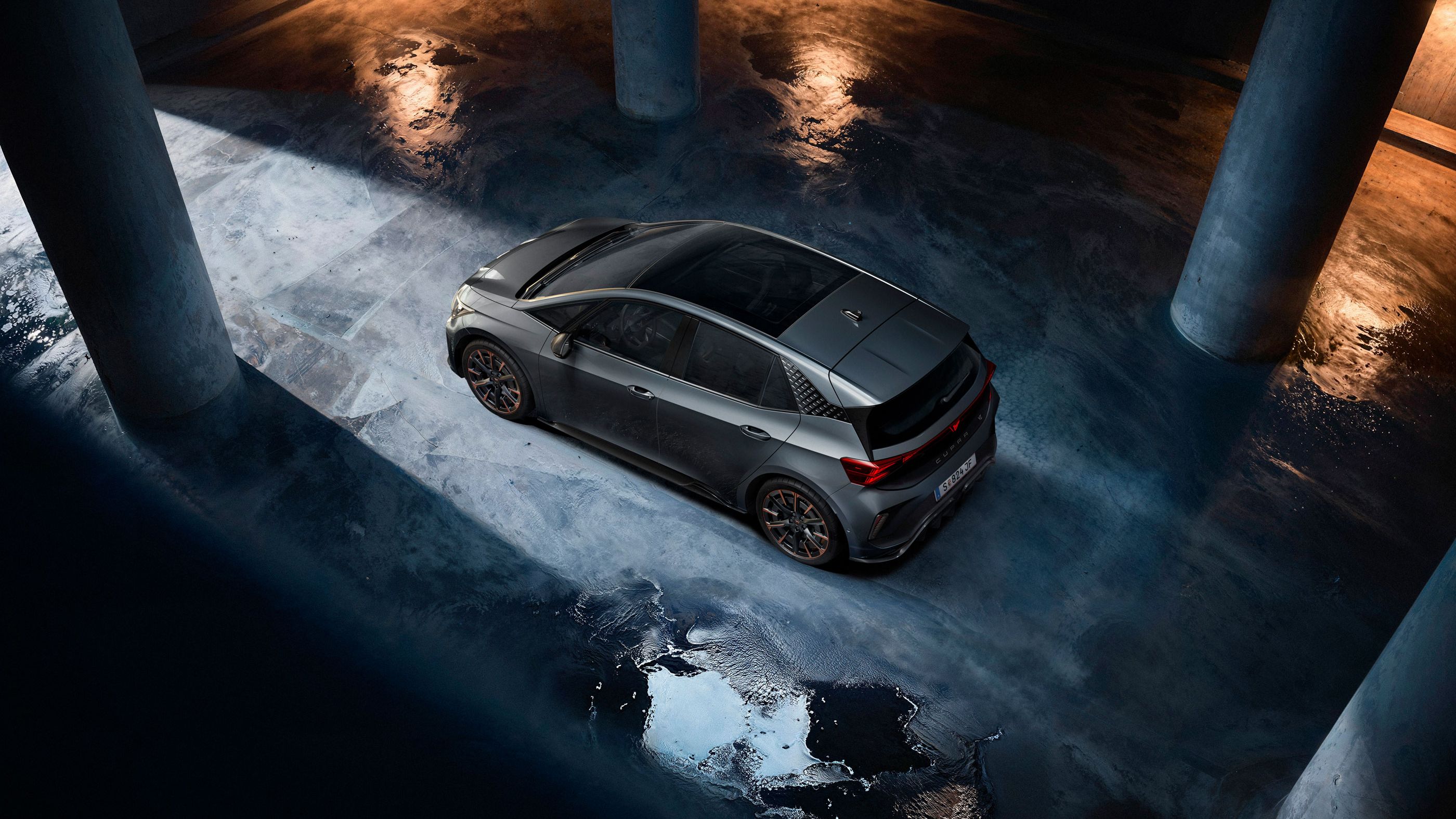 Ein grauer CUPRA Born steht in einer dunklen Tiefgarage. Das Bild ist aus der Vogelperspektive aufgenommen und zeigt Lichterscheinungen auf dem nassen Betonboden. Die Szenerie ist durch indirektes Licht beleuchtet, das eine dramatische Atmosphäre schafft.