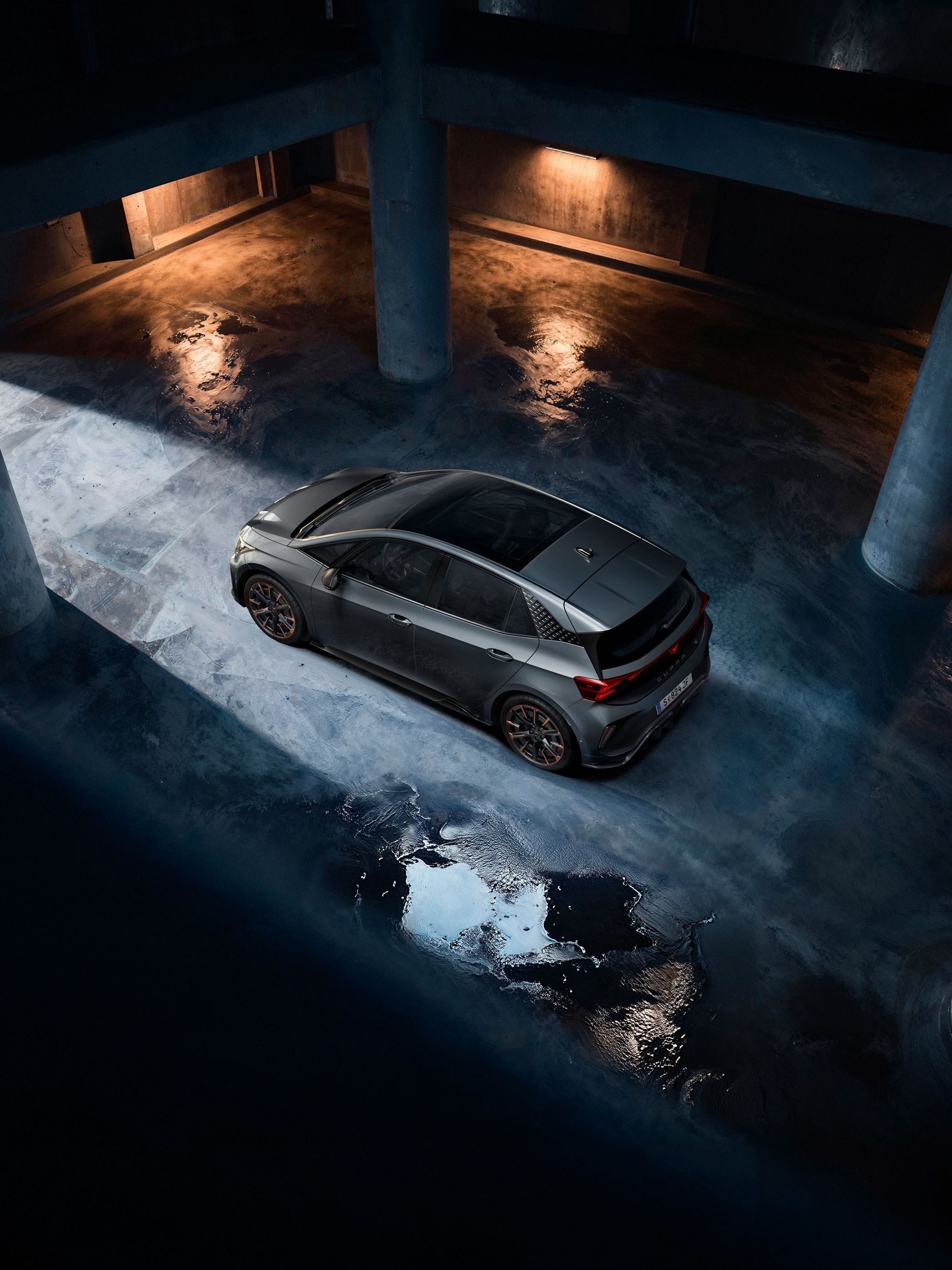 Ein grauer CUPRA Born steht in einer dunklen Tiefgarage. Das Bild ist aus der Vogelperspektive aufgenommen und zeigt Lichterscheinungen auf dem nassen Betonboden. Die Szenerie ist durch indirektes Licht beleuchtet, das eine dramatische Atmosphäre schafft.